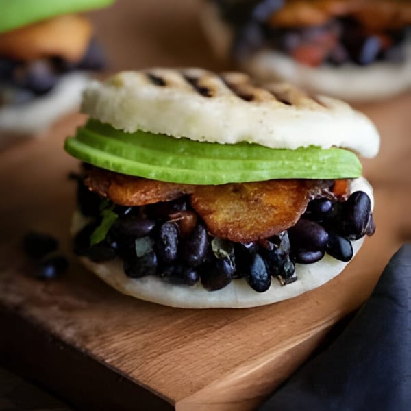 A Sabrosa Venezuela arepa filled with black beans, fried plantains, and sliced avocado on a wooden board.