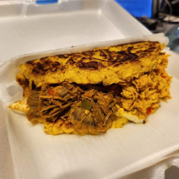 Close-up of a Sabrosa Venezuela cachapa, a golden corn pancake filled with shredded meat, in a white takeout container.