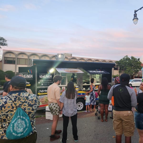 People gather around the Sabrosa Venezuela food truck at dusk, with a building in the background.