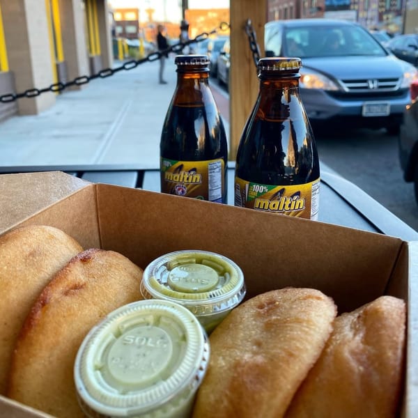 A takeout box of golden-fried empanadas with green sauces and two bottles of Maltin on a table.